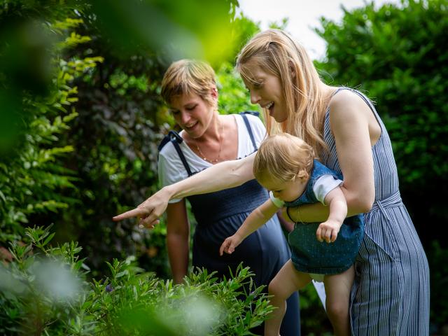 Two women and a small child in a garden, smiling and pointing at something in the shrubs